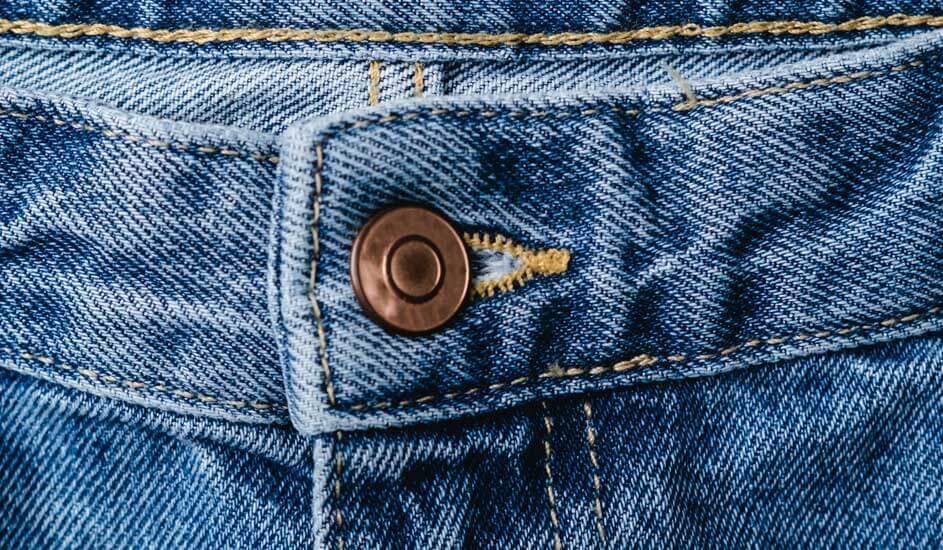 A close-up of blue denim jeans shows the waistband, a metal button and yellow stitching. The texture and details of the fabric are clearly visible. No brand shown.
