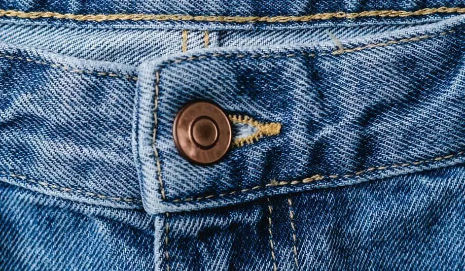 A close-up of blue denim jeans shows the waistband, a metal button and yellow stitching. The texture and details of the fabric are clearly visible. No brand shown.