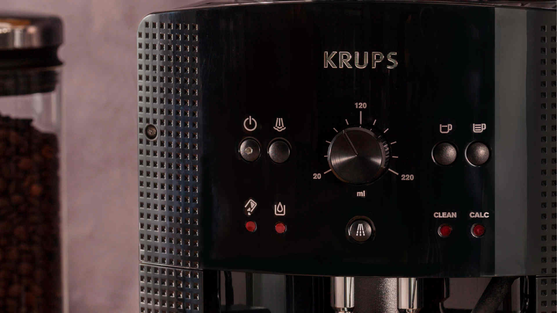 Zwarte Krups koffiemachine met knoppen, een draaiknop voor de hoeveelheid en rode indicatielampjes. Koffiebonen in een pot op de achtergrond.