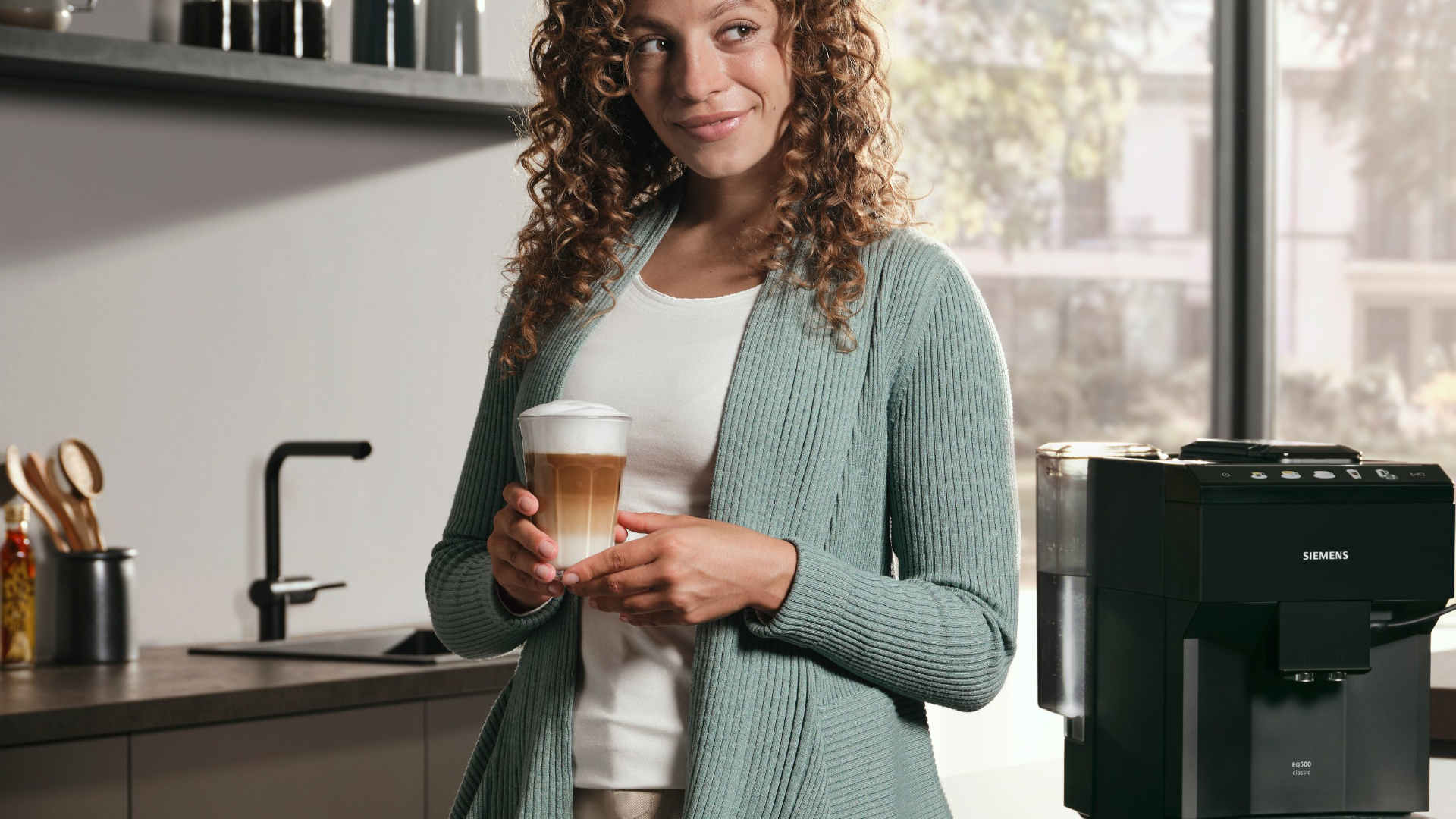Vrouw houdt een glas latte in de keuken, naast een Siemens koffiemachine en een gootsteen.