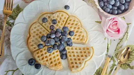Wafel op wit bord met blauwe bessen, naast een kom bessen, roos, mes, lepel en vork op een bloemig doek.