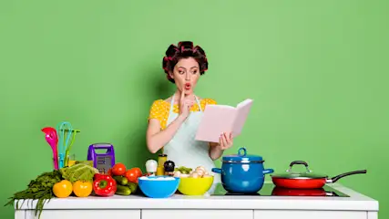 Vrouw met een kookboek, omringd door groenten, fruit, keukengerei, een blauwe pan en een rode koekenpan op een witte tafel tegen een groene achtergrond.