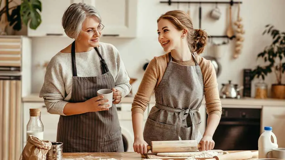 Twee vrouwen in schorten bakken in een keuken. Een vrouw rolt deeg uit, de andere houdt een mok vast.