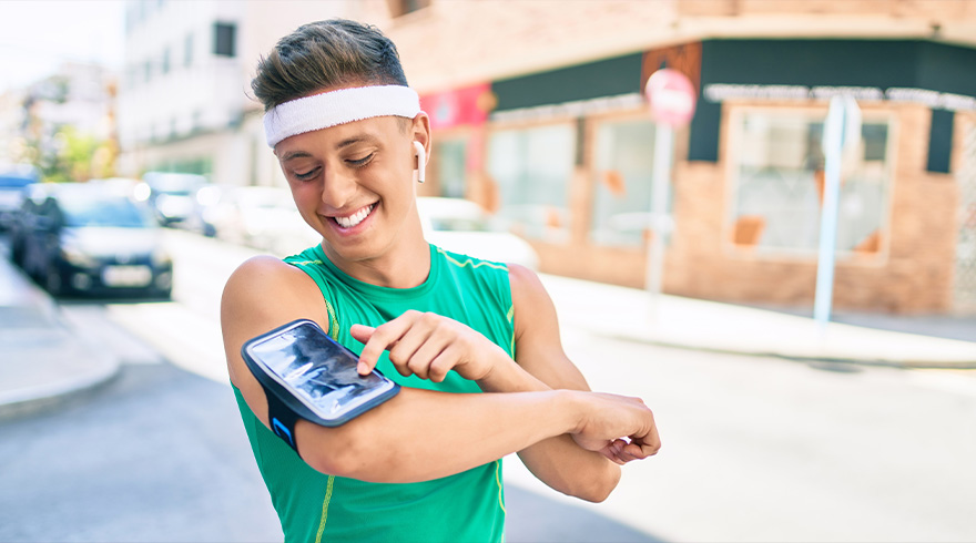 Een man glimlacht buiten, draagt een groen hemd en een witte hoofdband, en gebruikt een telefoon in een armhouder.