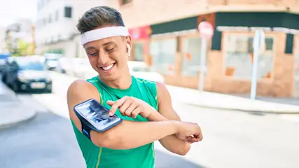 Een man glimlacht buiten, draagt een groen hemd en een witte hoofdband, en gebruikt een telefoon in een armhouder.