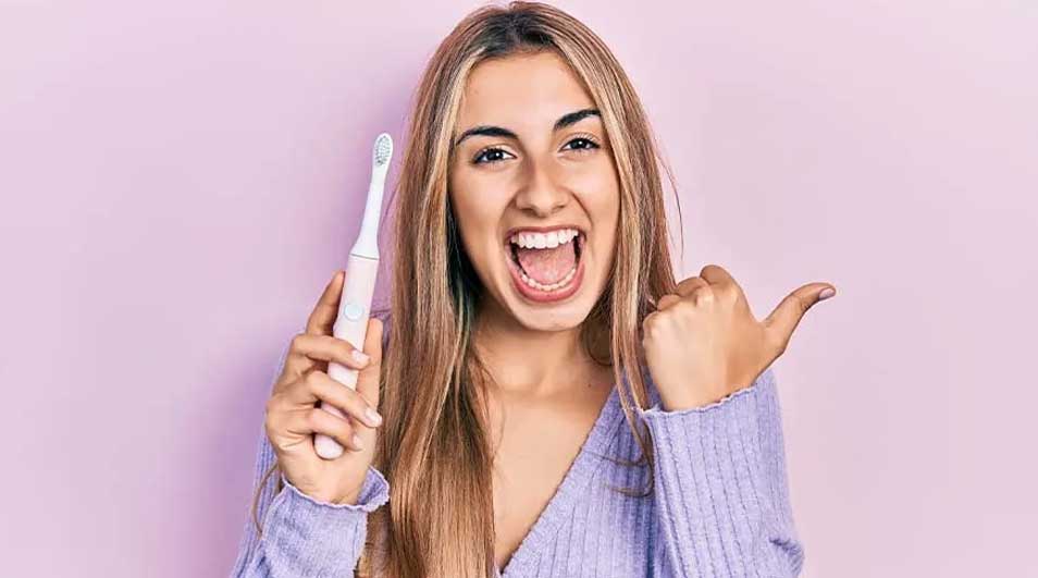 Een vrouw lacht en houdt een witte elektrische tandenborstel vast terwijl ze naar rechts wijst tegen een paarse achtergrond.