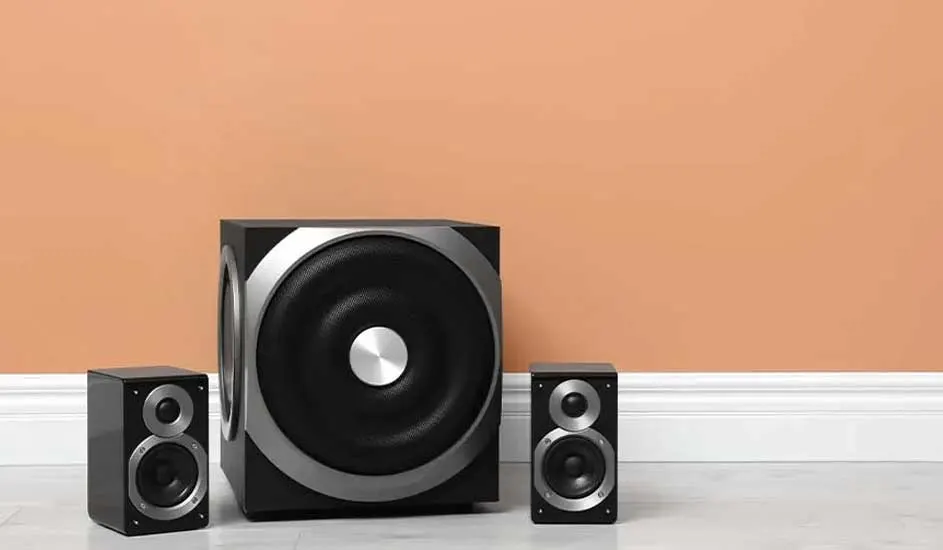 Black subwoofer with two compact speakers, positioned on a marble floor. Background features a peach-coloured wall, sleek and modern.