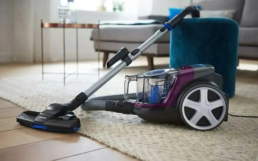 A modern Philips vacuum cleaner on wooden flooring, partly on a light rug, beside a teal pouffe and grey sofa in a tidy living room.