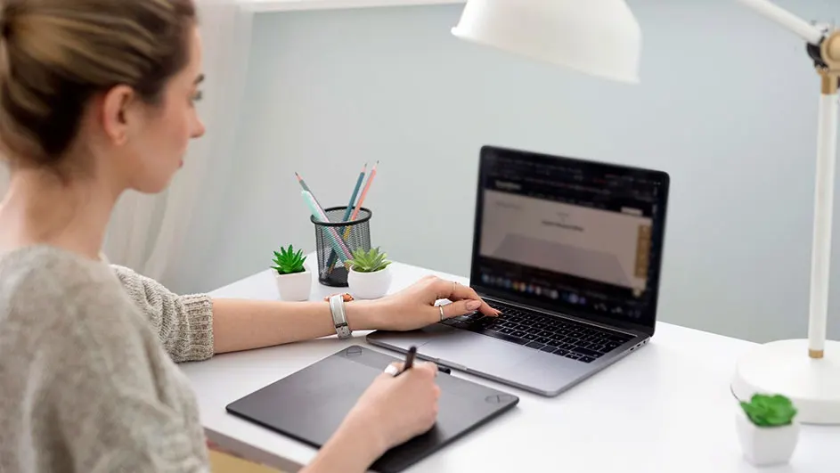 A person uses a laptop and graphic tablet on a white desk. A lamp, plants, and pens are nearby.