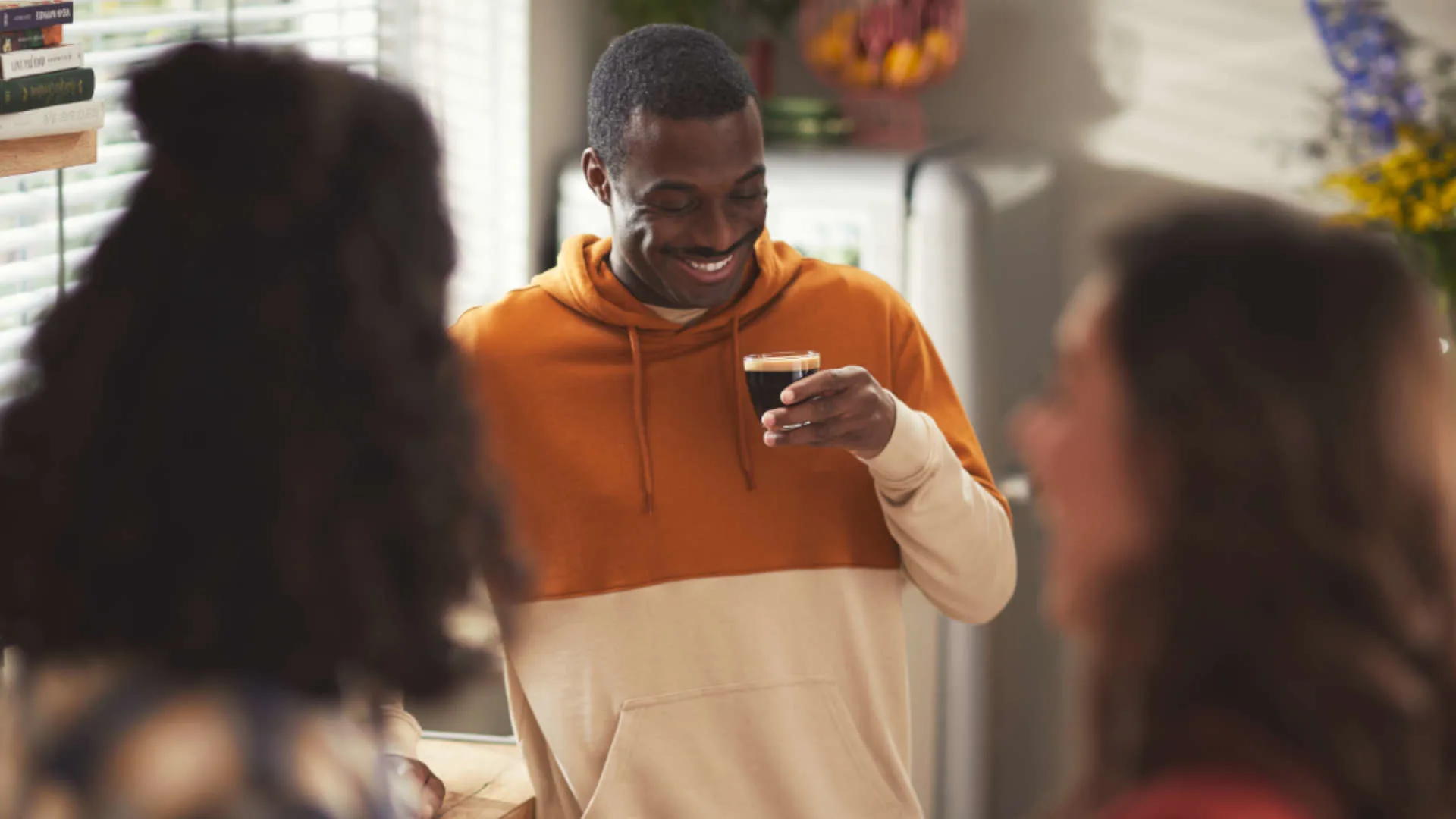 Een man in een oranje hoodie lacht en houdt een glas koffie vast, met mensen en een koelkast op de achtergrond.