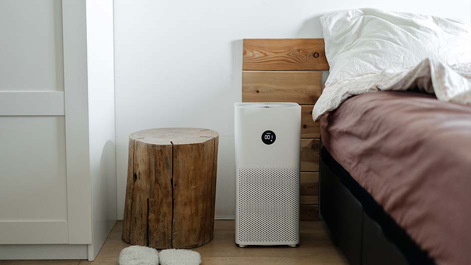 Een slaapkamer met een houten nachtkastje, een witte luchtreiniger, een bed met bruin beddengoed en een wit kussen.