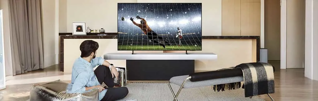 Een man zit op de vloer en kijkt naar een groot wandmontage-tv met voetbalwedstrijd. Modern interieur met houten meubels, grijze gordijnen, licht. Minimalistische, stijlvolle woonkamer setup.