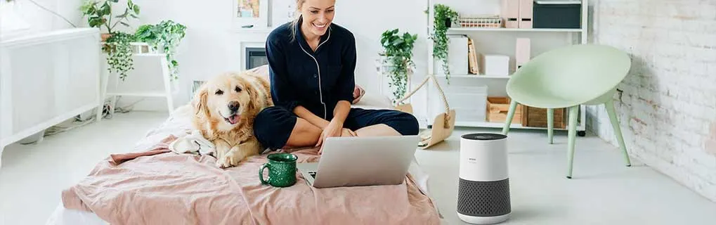 Vrouw zit lachend op bed met laptop naast haar hond, in lichte kamer met planten, terwijl een witte luchtreiniger stil op de vloer staat.