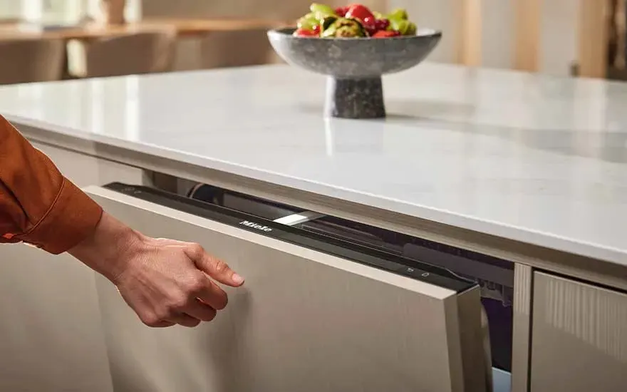 Een persoon opent een ingebouwde Miele vaatwasser in een moderne keuken met licht werkblad. Op het aanrecht staat een schaal met vers fruit.