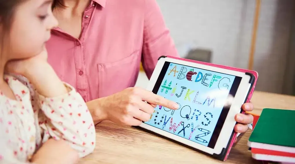 Een volwassene wijst op een Apple iPad naar kleurrijke letters van het alfabet, terwijl een kind meekijkt aan een houten tafel met boeken.