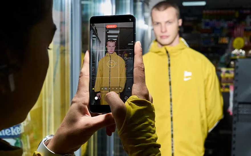 Someone films a man in a yellow jacket inside a shop, the Samsung smartphone screen showing a video interface with framing grid and recording timer.