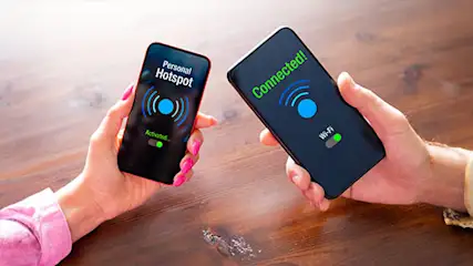 Two hands hold phones on a wood table. One phone displays "Personal Hotspot," the other "Connected!" Both show a blue wifi symbol.