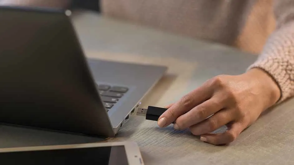Een hand steekt een zwarte USB-stick in een grijze laptop op een houten tafel. Een witte smartphone ligt ernaast.