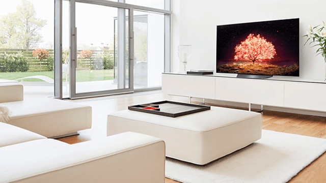 Woonkamer met witte bank, ottoman, tv-meubel en grote ramen. Een tv toont een rode boom.