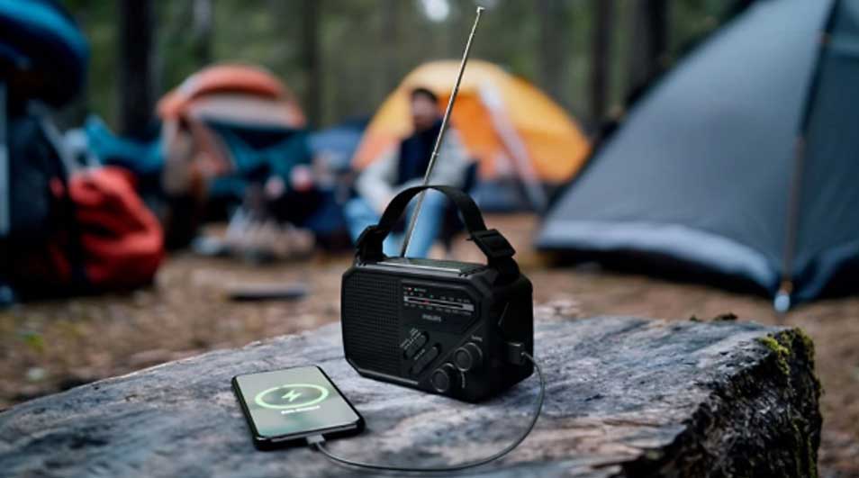 Op een boskamp staat een draagbare zwarte radio op een boomstam, verbonden met een smartphone, terwijl tenten vaag oplichten.