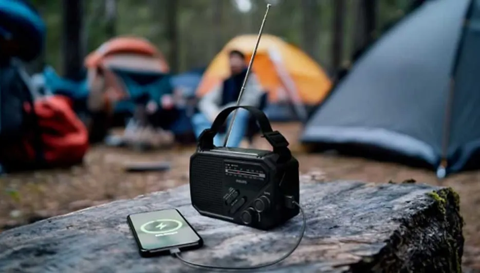 Op een boskamp staat een draagbare zwarte radio op een boomstam, verbonden met een smartphone, terwijl tenten vaag oplichten.