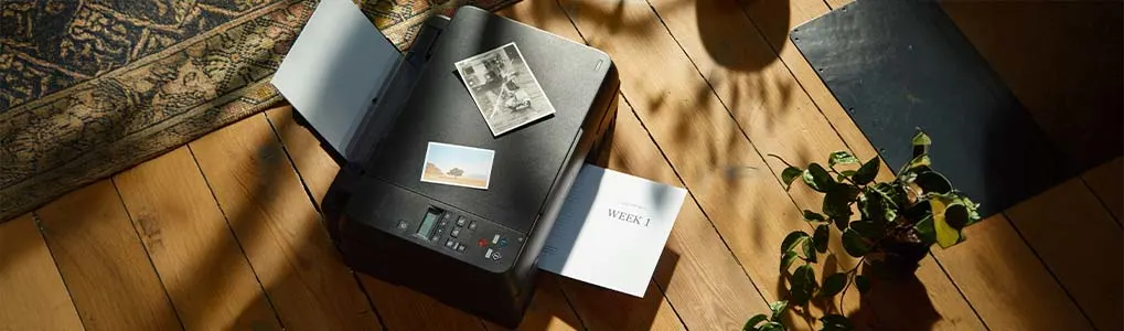 Een printer op een houten vloer print een vel papier; er liggen ook twee foto's bovenop.