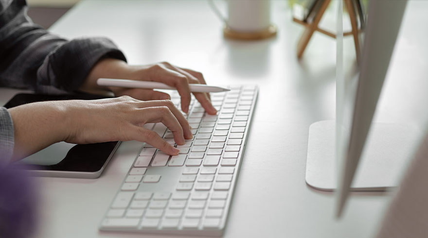 Twee handen typen op een wit toetsenbord naast een tablet en stylus op een witte tafel. Op de achtergrond staat een witte mok.