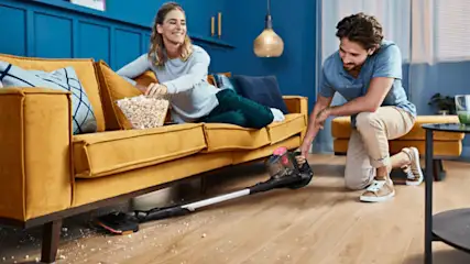 Een man stofzuigt onder een mosterdgele bank terwijl een vrouw met popcorn op de bank zit in een blauwe kamer.