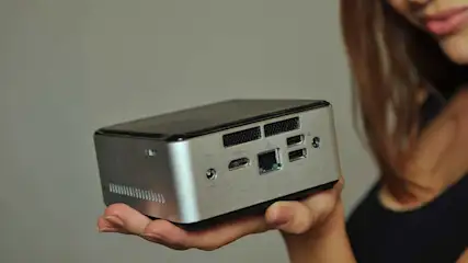 Een persoon houdt een kleine, zilverkleurige computer vast met poorten en ventilatieopeningen zichtbaar. De achtergrond is grijs.