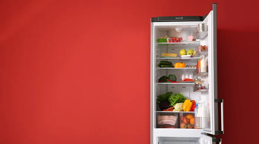 Een open witte koelkast gevuld met diverse groenten en fruit, staat tegen een effen rode achtergrond.