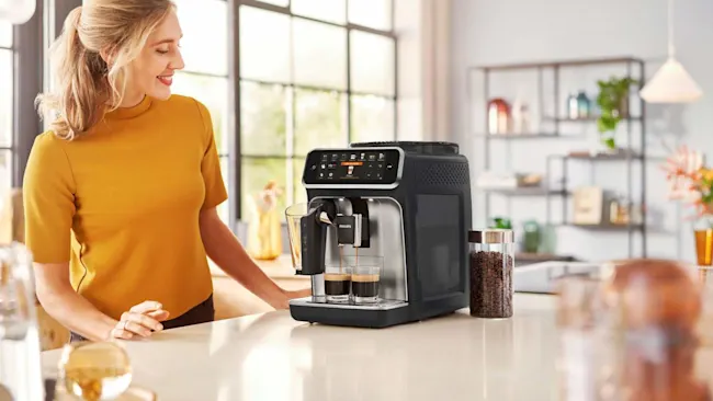 Een vrouw in een gele trui staat naast een espressomachine die twee kopjes koffie zet, met een pot koffiebonen ernaast.