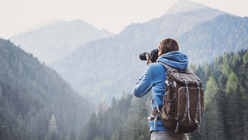 Een persoon met rugzak fotografeert met een camera in een bergachtig boslandschap.