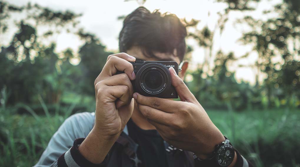 Een persoon houdt een zwarte camera vast met beide handen tegen een groene, wazige achtergrond.
