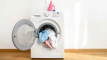 Een witte wasmachine met een open deur, gevuld met blauwe, roze en witte kleding, staat op een houten vloer tegen een witte muur.