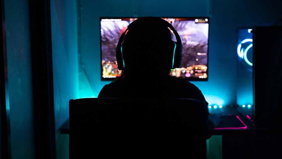 Donkere kamer met persoon met koptelefoon voor een computerscherm. Blauwe verlichting accentueert de randen van de schermen en de kamer.