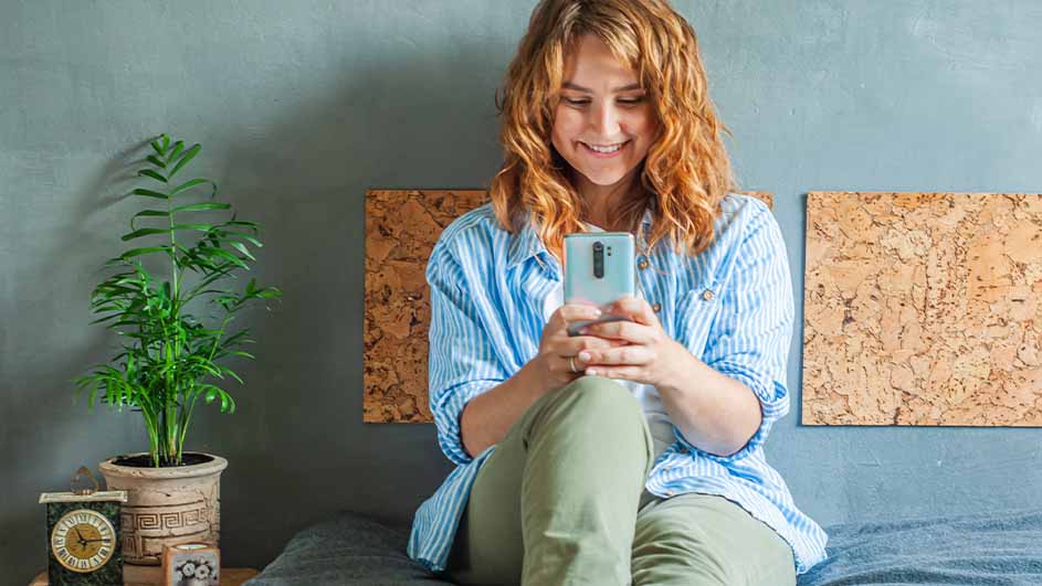 Vrouw zit op een bed met groene broek en gestreept blauw overhemd, kijkend naar een blauwe mobiele telefoon. Kamerplant en klok zichtbaar.