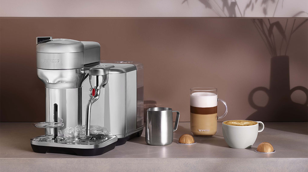 Een zilverkleurig koffiezetapparaat naast kopjes koffie en capsules op een aanrecht.