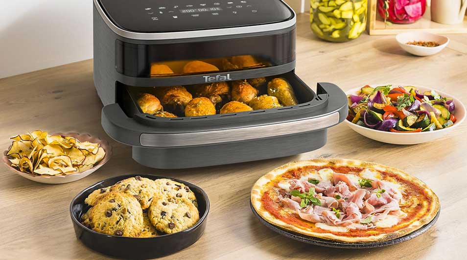 A Tefal air fryer sits on the table with chicken, biscuits, pizza, chips and a bowl of grilled vegetables around it.