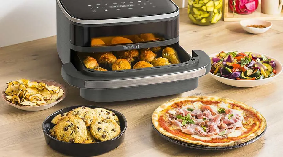 A Tefal air fryer sits on the table with chicken, biscuits, pizza, chips and a bowl of grilled vegetables around it.