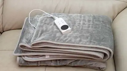 Een grijze, opgevouwen elektrische deken met een witte afstandsbediening ligt op een beige bank.