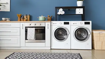 Witte kasten, een oven en wasmachines tegen een blauwe muur. Een zwart-wit vloerkleed ligt op de vloer.