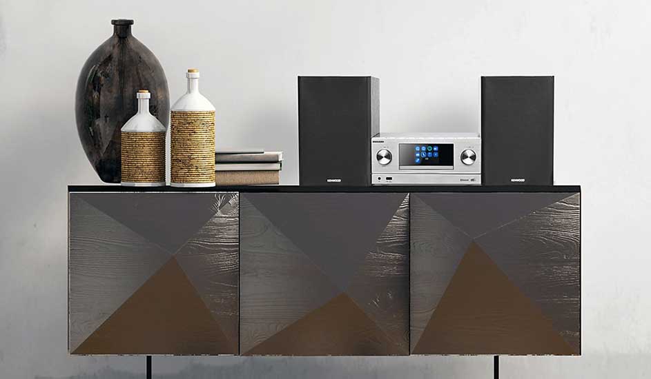 Op een modern dressoir staan twee zwarte speakers, een zilveren stereo, flessen en boeken, naast een grote donkere vaas.