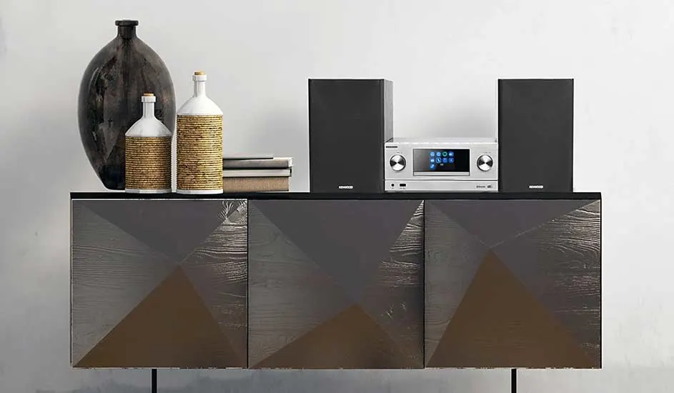 Op een modern dressoir staan twee zwarte speakers, een zilveren stereo, flessen en boeken, naast een grote donkere vaas.