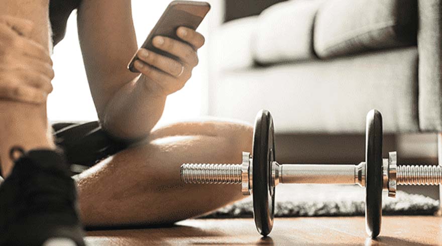 Een persoon zit op de vloer met een telefoon in de hand, naast een halter. Een bank is zichtbaar op de achtergrond.