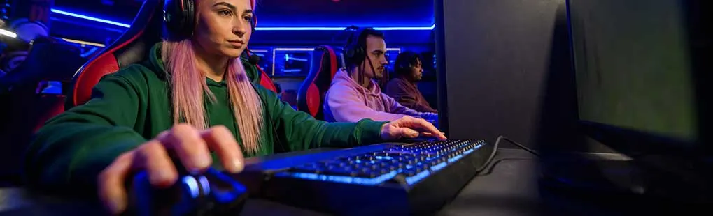Gamers met headsets spelen gefocust op pc’s in een neonverlichte esports-ruimte; vrouw vooraan met muis en toetsenbord. Geen zichtbaar merk.