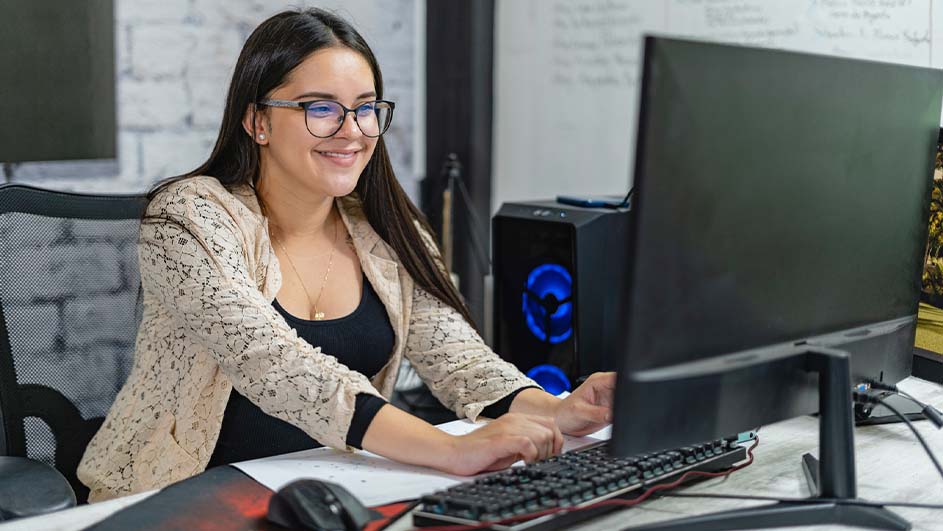 Vrouw zit aan een bureau met een computer, toetsenbord en muis. Achter haar staat een computer met blauwe verlichting en een witte bakstenen muur.