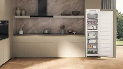 Een keuken met beige kasten, een marmeren achterwand en een openstaande witte diepvriezer.