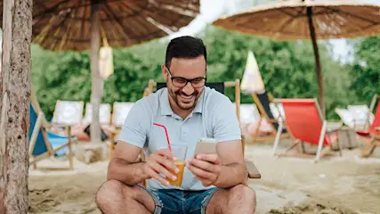 Een man met een bril zit op een strandstoel met een drankje en een telefoon, met rieten parasols en stoelen op de achtergrond.