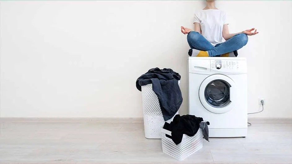 Person in jeans sits in lotus pose on a white washing machine. Two laundry baskets hold dark clothes.