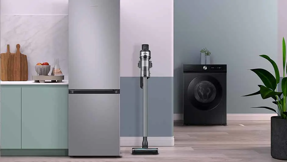 Koelkast, stofzuiger en wasmachine staan in een kamer. Er staan een houten snijplank en een kom met fruit op een kast.